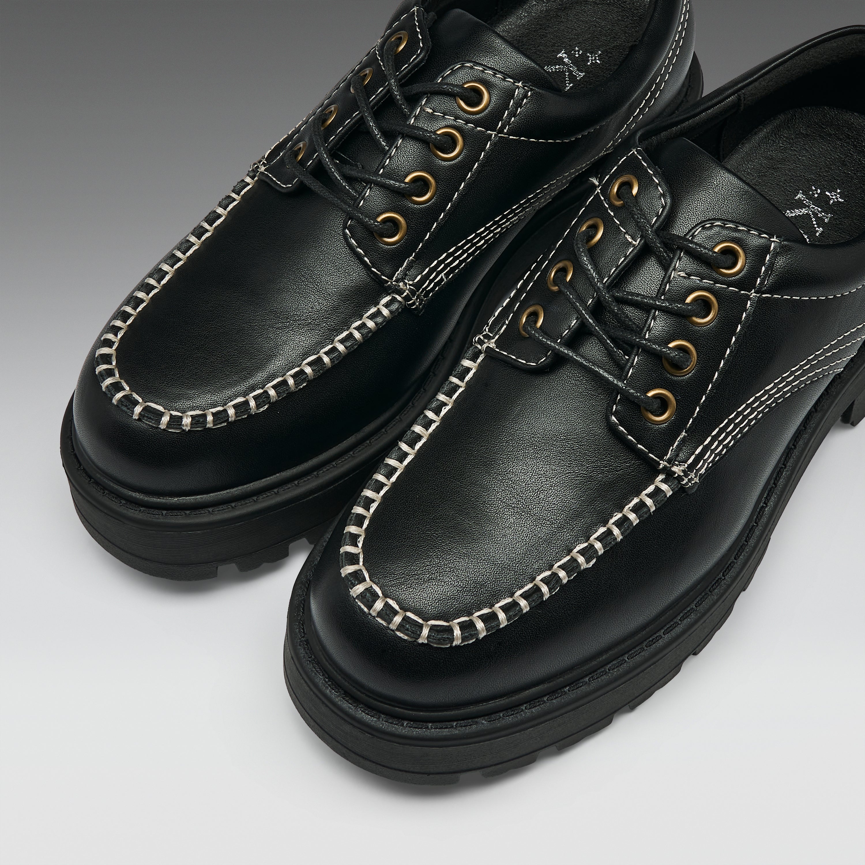 A pair of black leather lace-up shoes with distinctive white contrast stitching along the toe, gold-tone metal eyelets, and thick black soles, shown on a light gray gradient background.