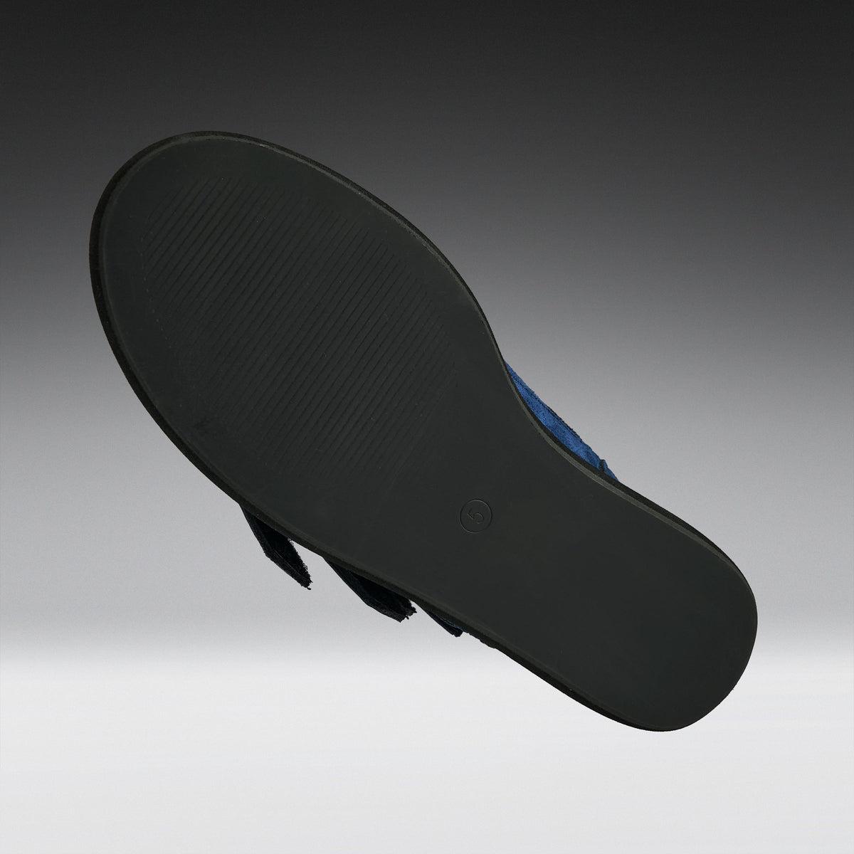 The black outsole of a flat sandal, shown floating against a dark gray to light gray gradient background. A small portion of the blue upper is visible on the right side.