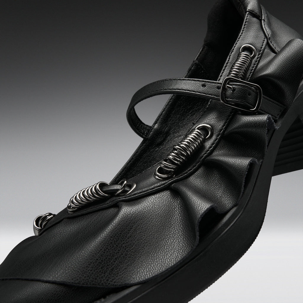 A close-up studio shot of a black leather Mary Jane-style shoe with multiple spring-loaded metal loops and a buckle strap, set against a dark gray gradient background.