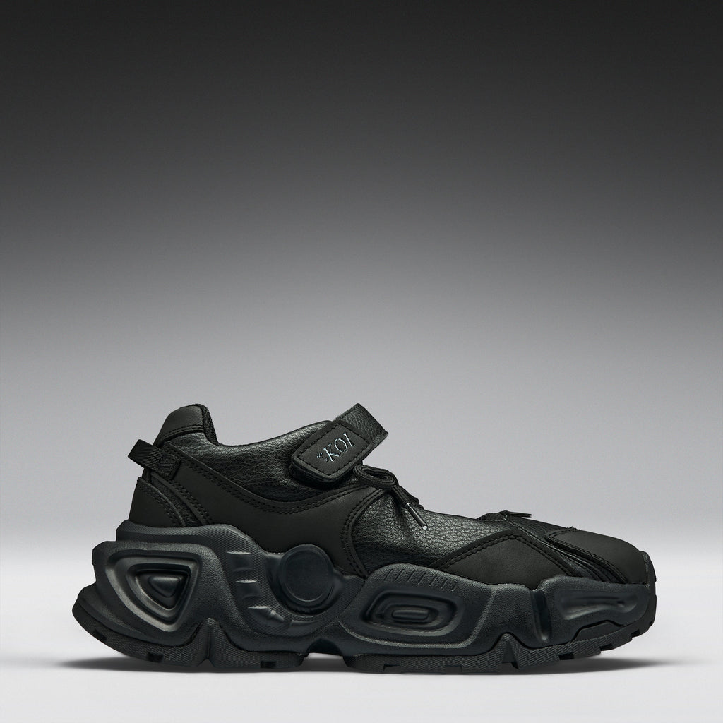 A black chunky platform sneaker with a hook-and-loop strap closure, shown from a side profile against a dark gray gradient studio background.
