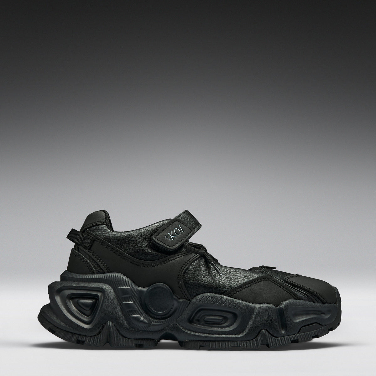 A black chunky platform sneaker with a hook-and-loop strap closure, shown from a side profile against a dark gray gradient studio background.
