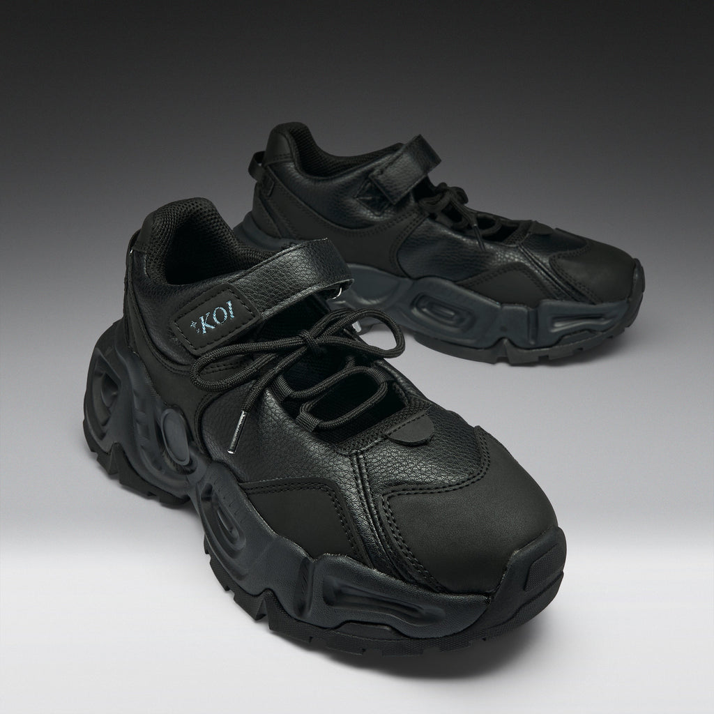 A pair of chunky black sneakers with a textured leather upper, thick sculpted black soles, and hook-and-loop strap closures, displayed on a dark-to-light gray gradient studio background.