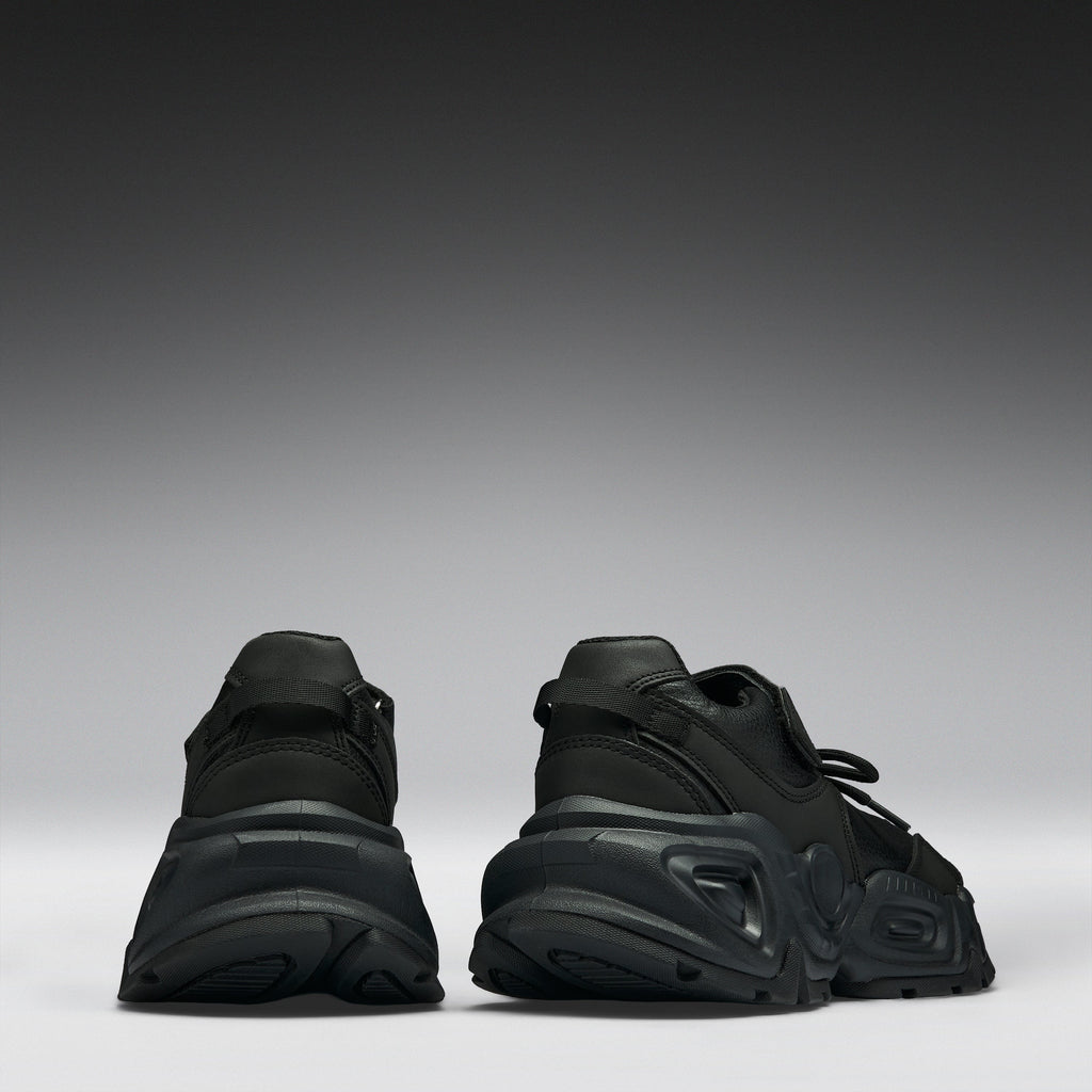 A pair of black chunky platform sneakers shown from the back, positioned side-by-side against a dark gray to light gray gradient studio background.