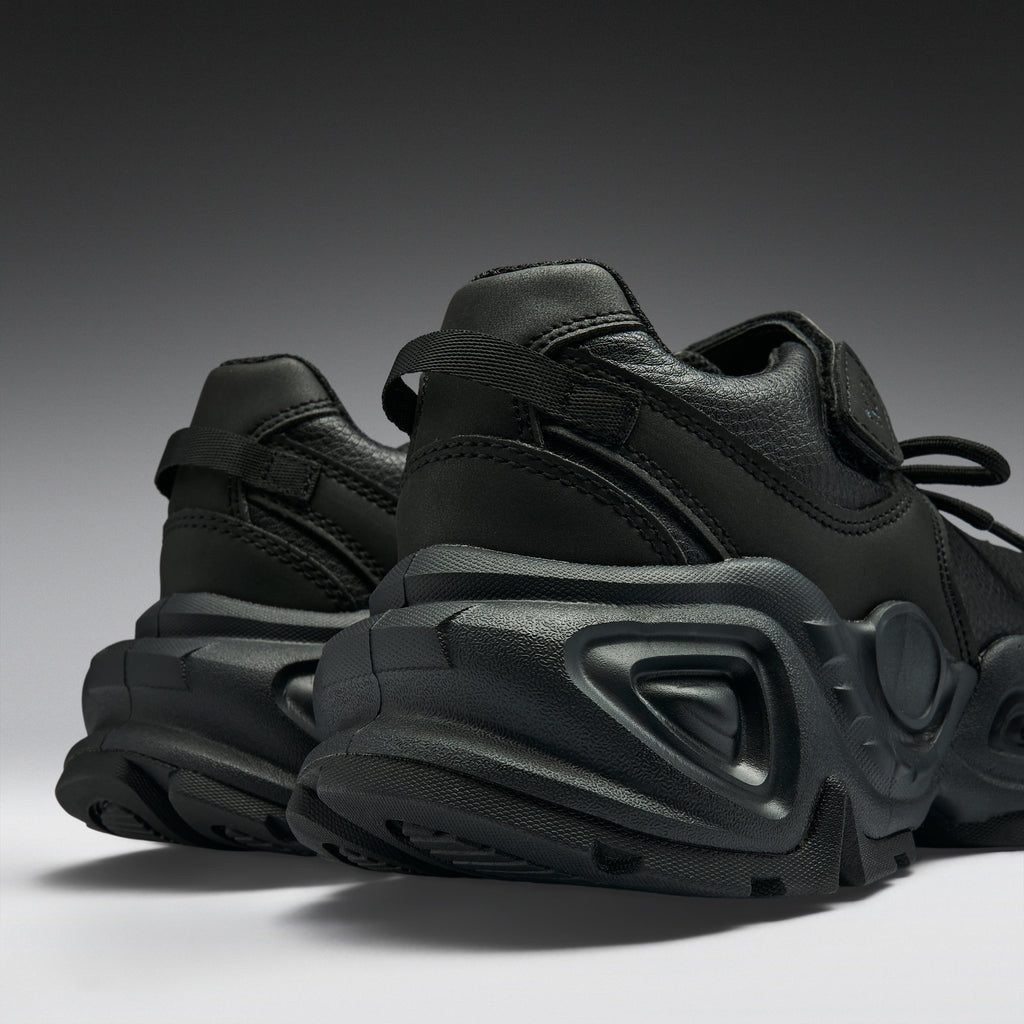 A pair of black chunky sneakers with a thick sculpted sole, shown from a low angle against a dark gray gradient background.