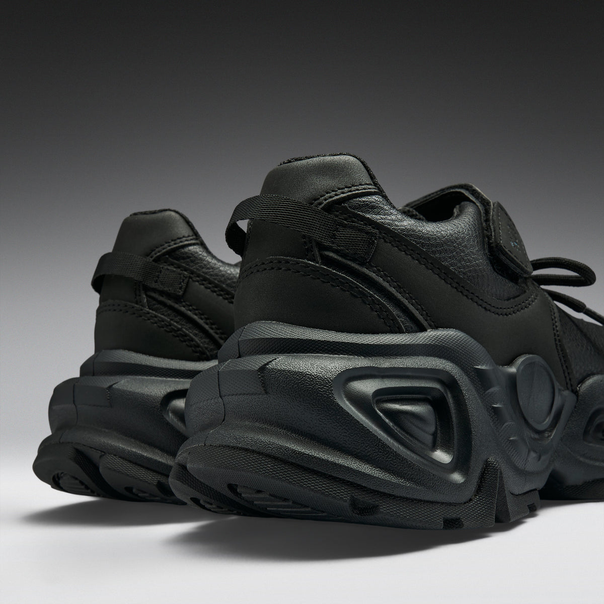 A pair of black chunky sneakers with a thick sculpted sole, shown from a low angle against a dark gray gradient background.