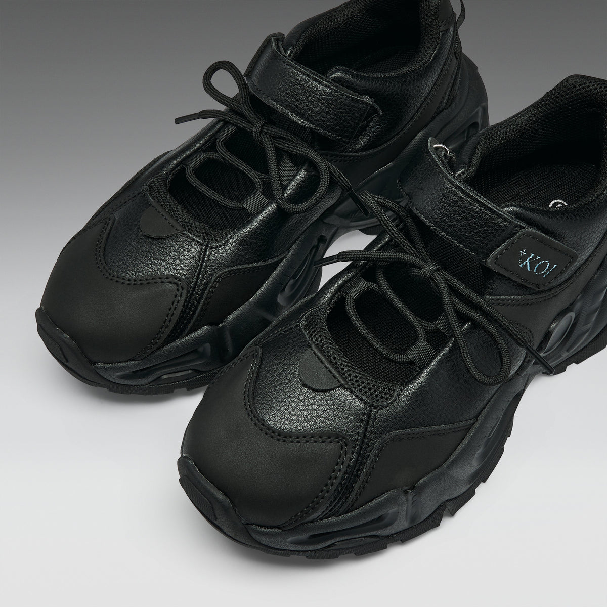 A pair of chunky black sneakers with a textured leather and mesh upper, featuring thick sculpted soles and hook-and-loop strap closures, shown on a neutral gray background.