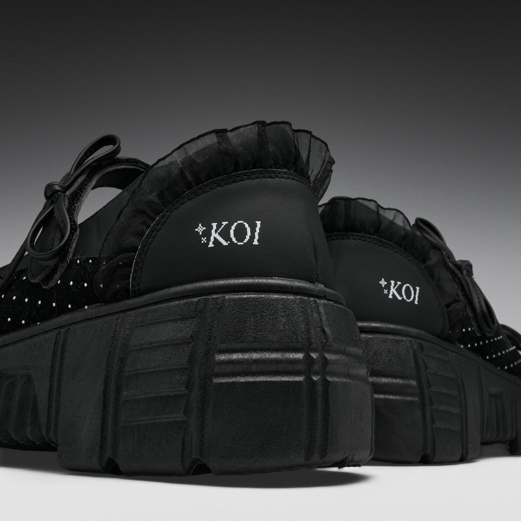 A pair of black platform sneakers with ruffled trim, studded details, and 'KOI' branding on the heel, shown against a dark gray gradient background.