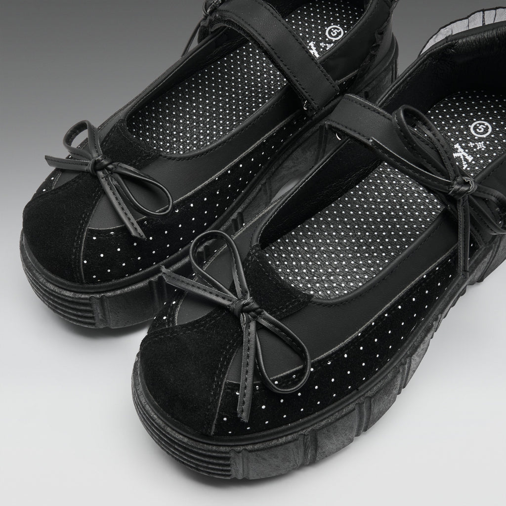 A pair of black platform Mary Jane shoes with suede toe caps, perforated details, and bow accents, shown on a neutral gray background.