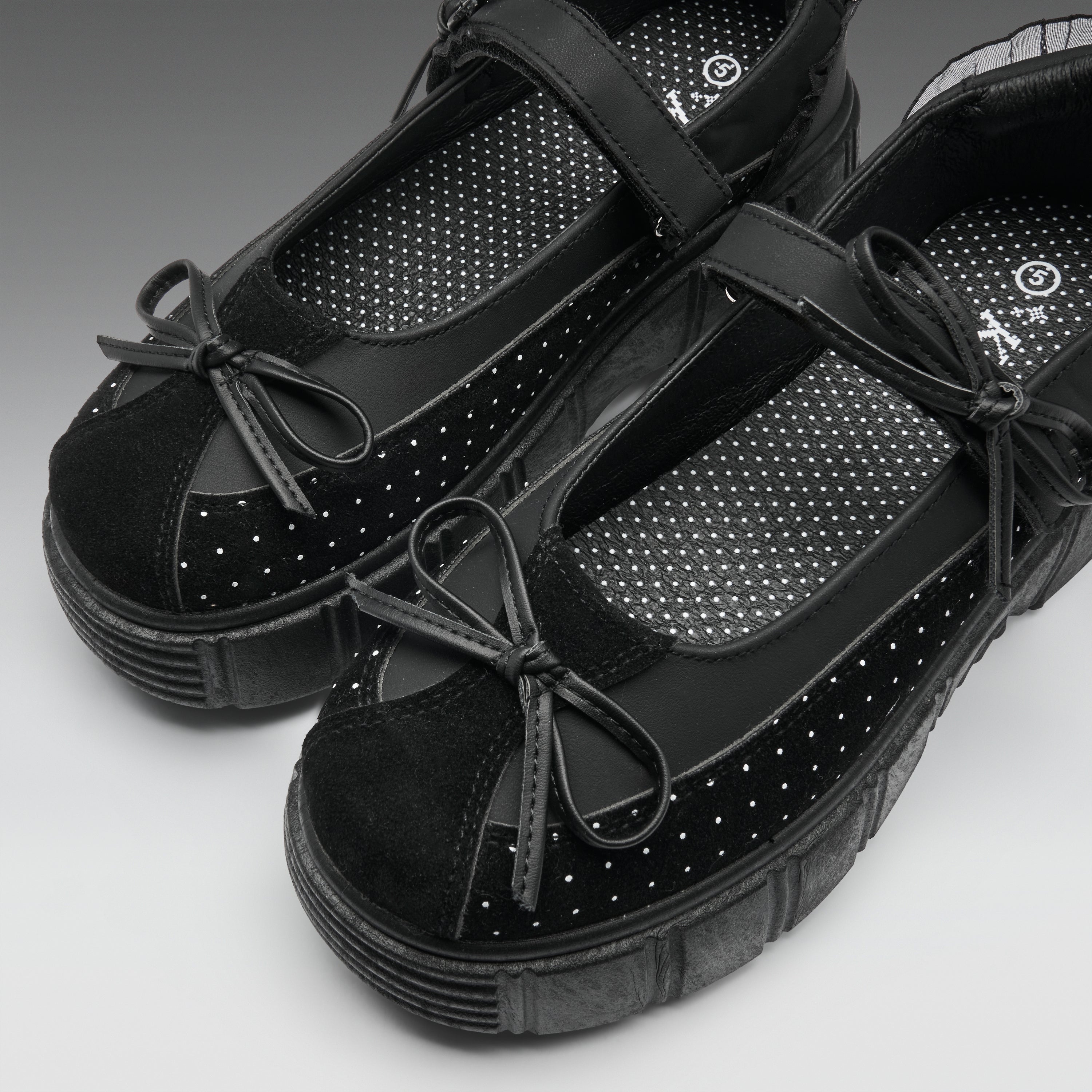 A pair of black platform Mary Jane shoes with suede toe caps, perforated details, and bow accents, shown on a neutral gray background.