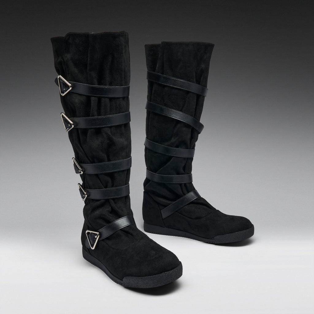 A pair of black knee-high boots with multiple wrap-around straps and silver-tone triangular buckle details, photographed on a dark-to-light gray studio background.