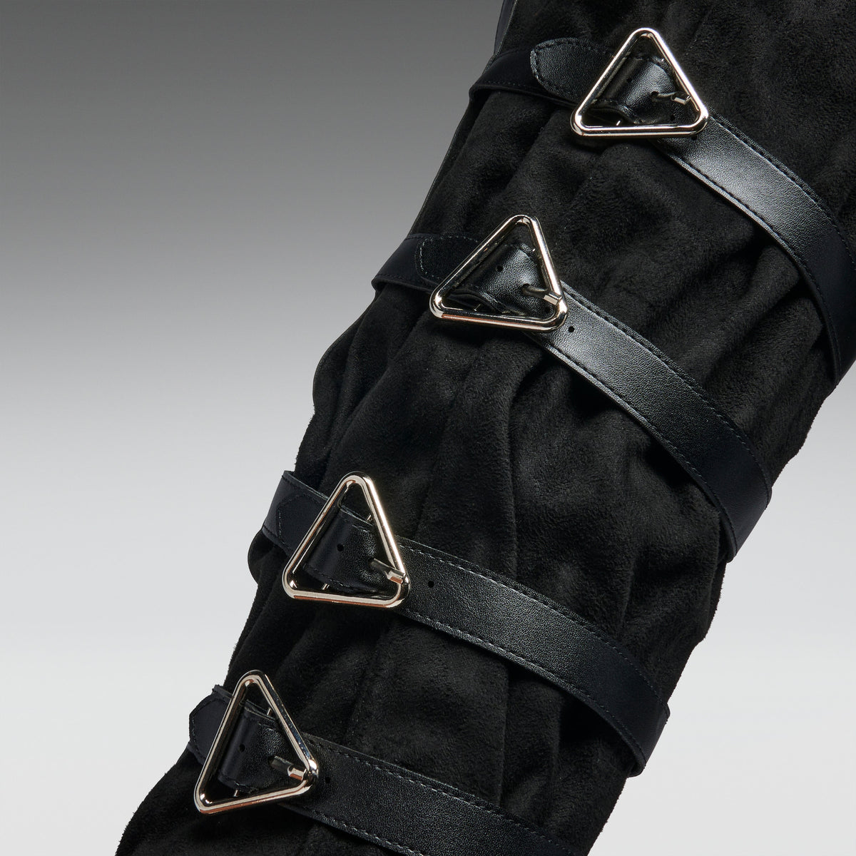 A close-up of a black textured boot or leg cover with multiple black straps and shiny silver triangular metal buckles, shown against a neutral gray gradient background.