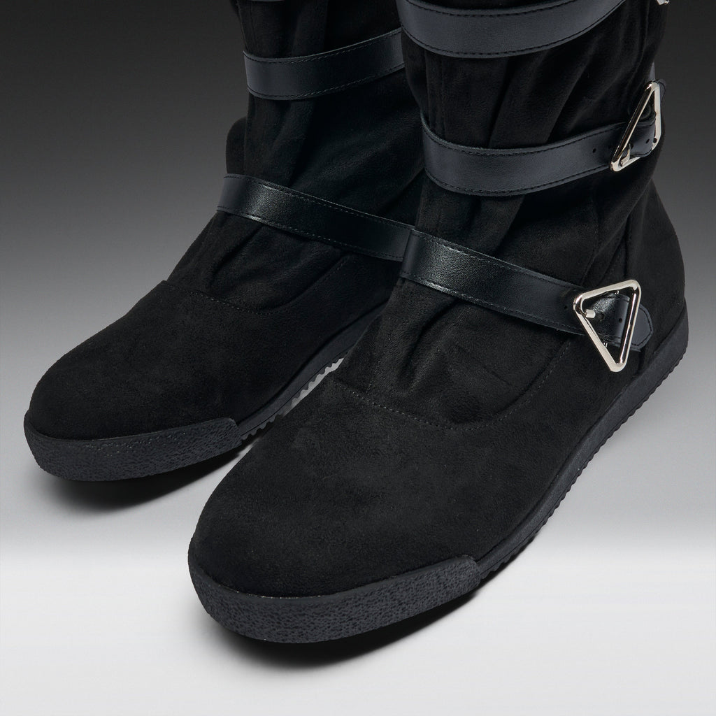 A pair of black suede and leather ankle boots with wide straps and silver triangular buckle details, shown on a neutral gray background.