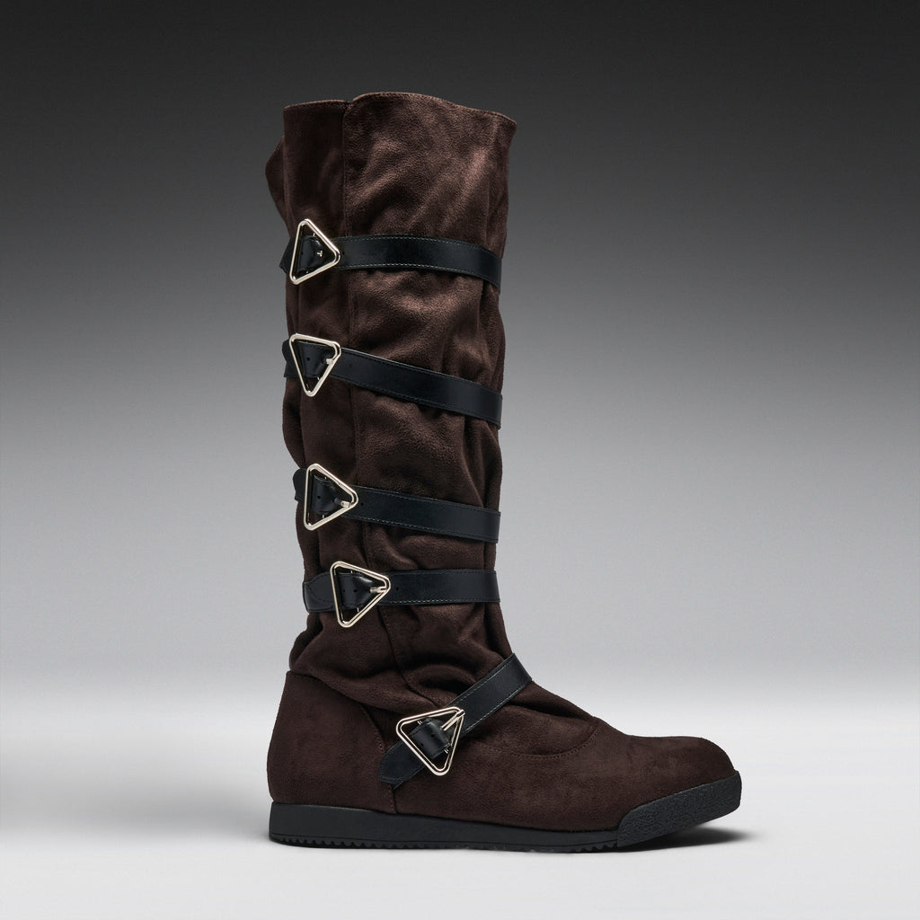 A dark brown suede knee-high boot with multiple black straps and silver-tone triangular metal hardware, shown in profile against a gray gradient studio background.