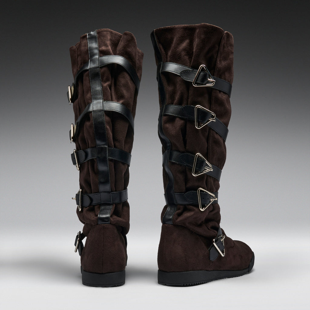 A pair of dark brown suede knee-high boots with multiple black faux-leather straps and silver-tone triangular buckle details, shown from the back on a gray gradient studio background.
