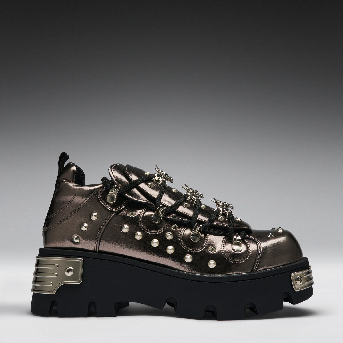 A metallic bronze high-top sneaker with black laces, silver studs, and a chunky black platform sole with silver metal accents, shown against a dark gray gradient background.