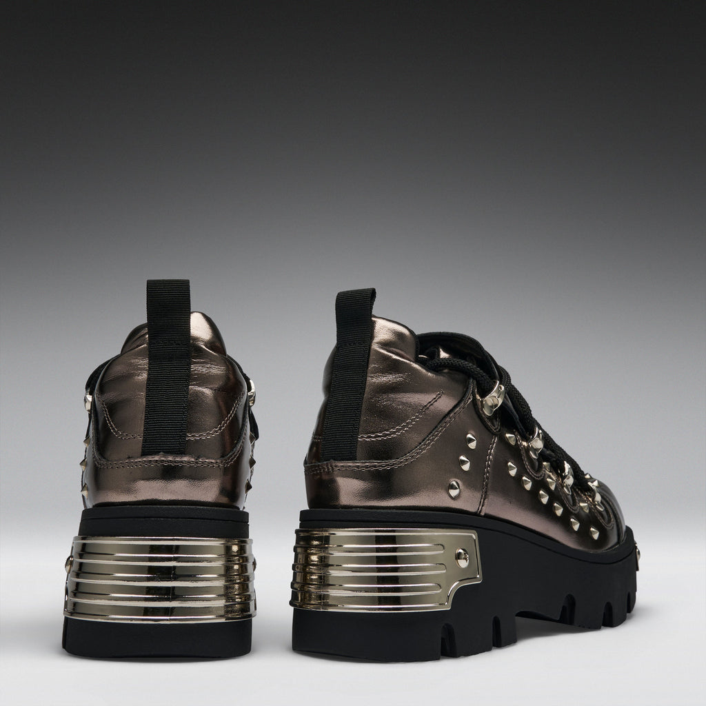 A pair of metallic bronze high-top platform sneakers with black soles, black laces, and silver studs, shown from the back and side against a dark gray gradient background.