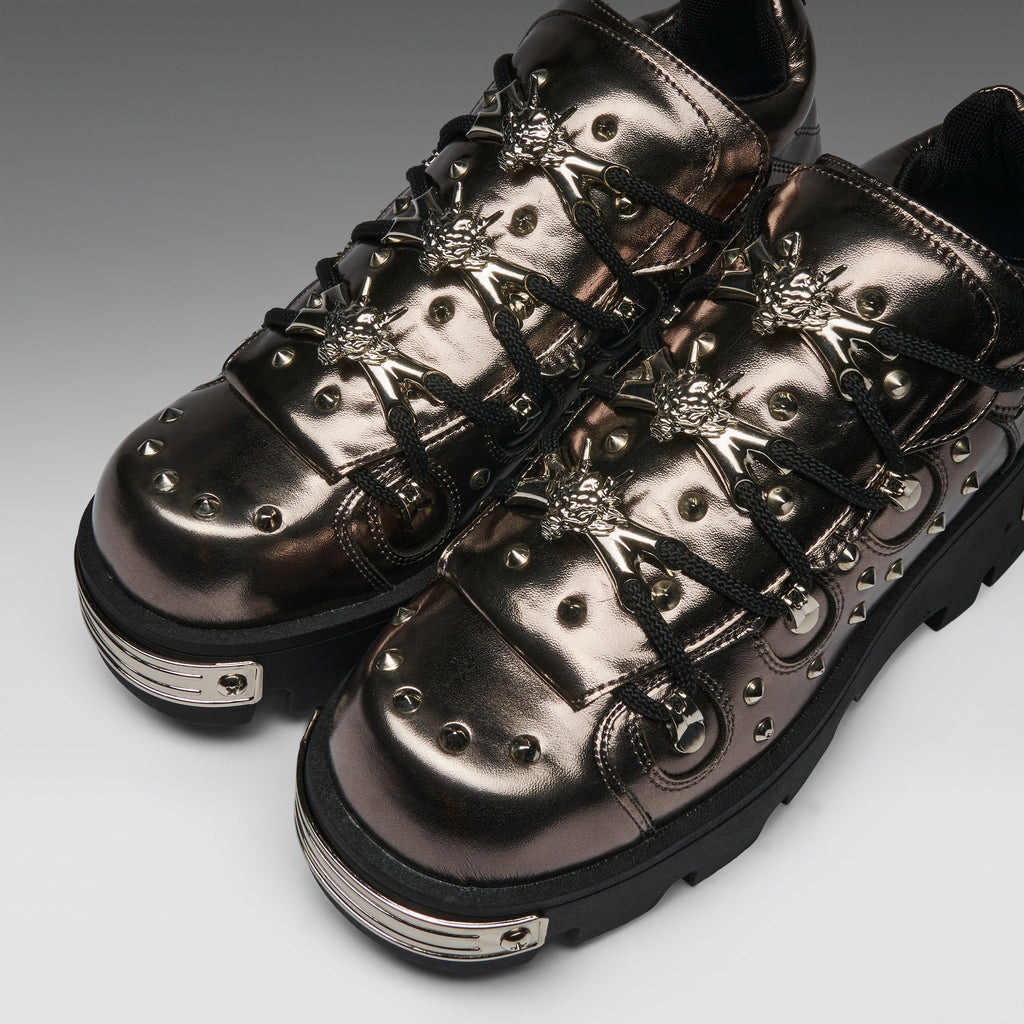 A pair of metallic bronze goth-style lace-up sneakers with silver studs, decorative metal hardware, and black laces, shown on a neutral gray background.