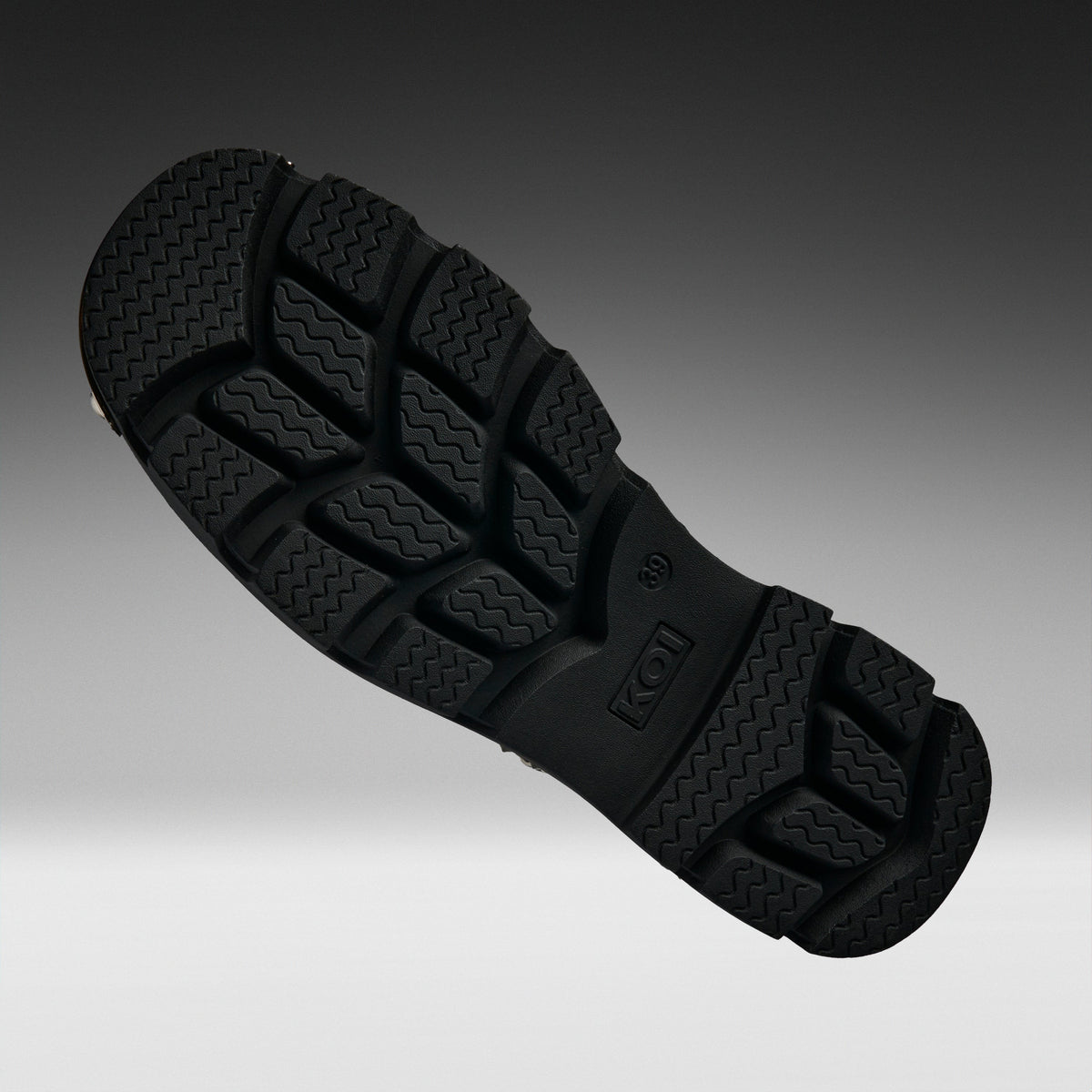 A close-up of a black rubber shoe sole with deep tread patterns, shown diagonally against a dark-to-light gray gradient background.
