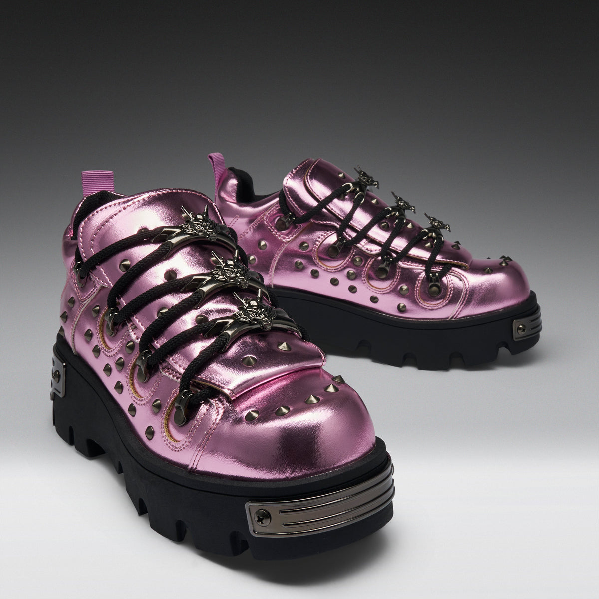 A pair of metallic pink platform sneakers with black lug soles, black decorative laces, and silver studs and hardware, photographed on a dark-to-light gray studio background.