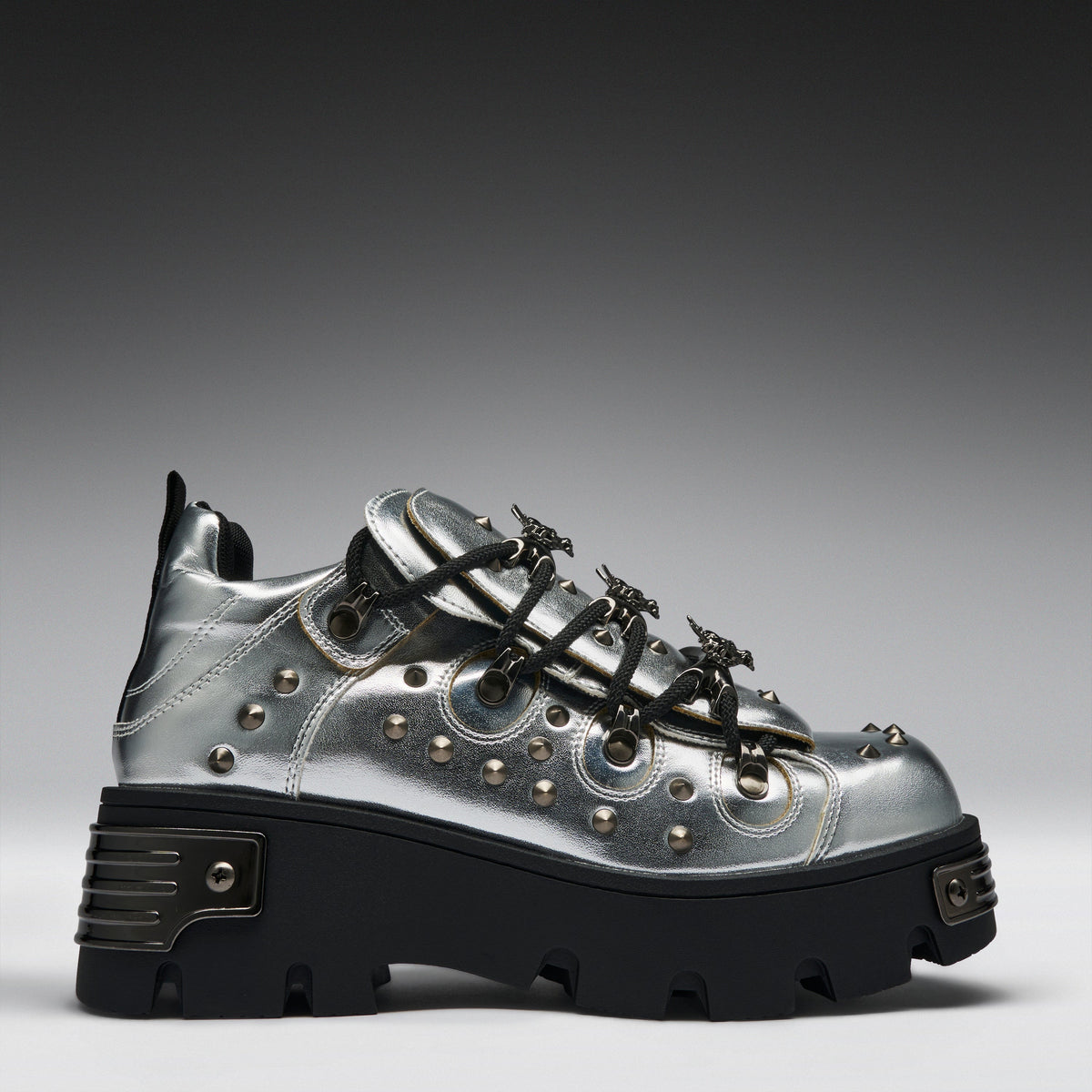 A metallic silver platform sneaker with black lug soles, black laces, and silver studs, shown from a side profile on a gray gradient background.