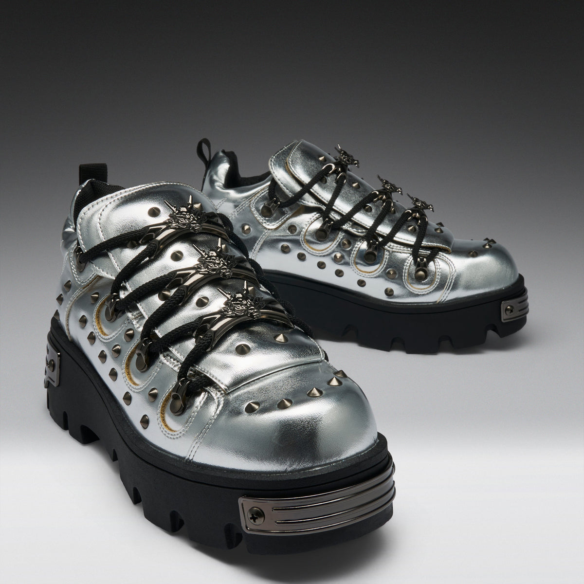 A pair of metallic silver high-top sneakers with black platform soles, black laces, and silver studs, displayed on a dark-to-light gray gradient studio background.