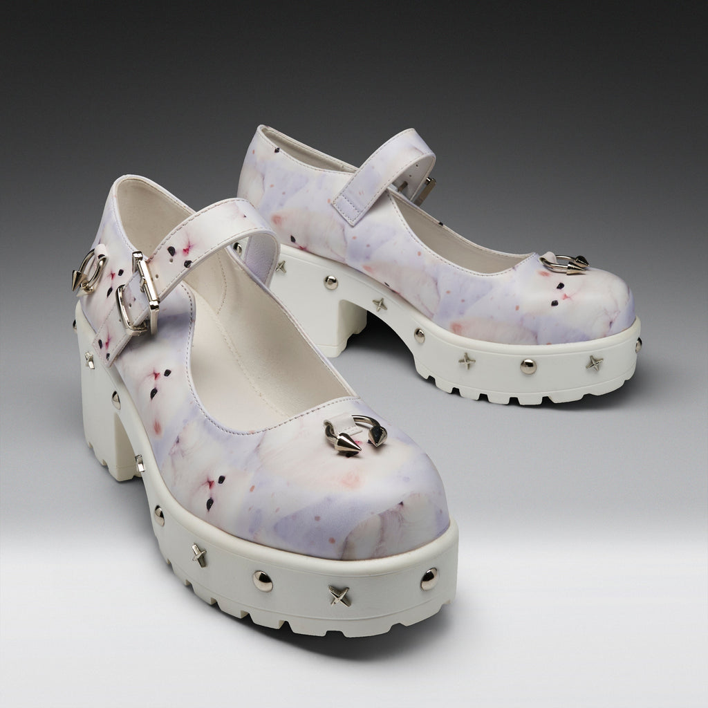 A pair of Mary Jane platform heels with a white and light purple watercolor pattern, decorated with silver buckles, spikes, and a metallic bow on the toe, set against a dark gray gradient background.
