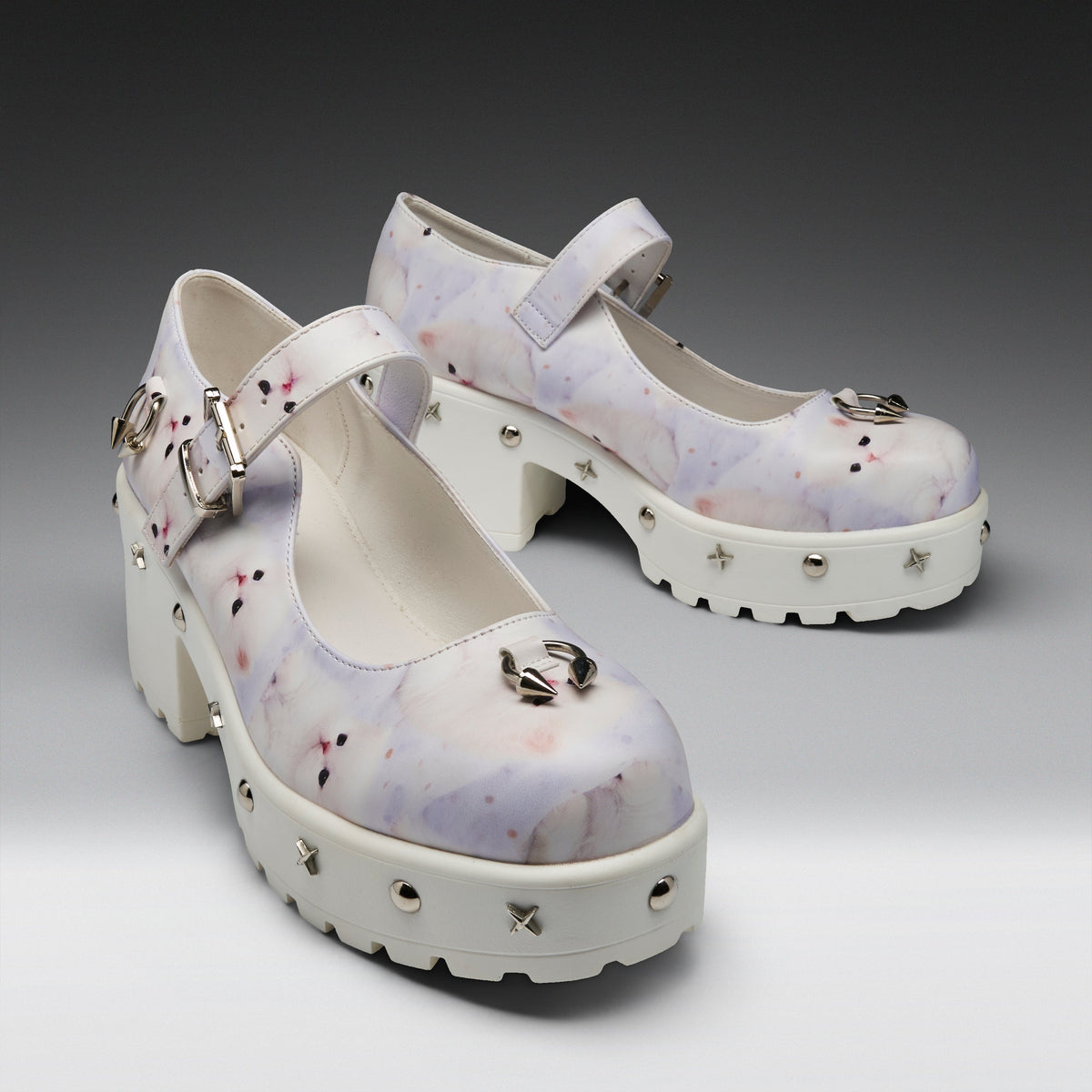 A pair of Mary Jane platform heels with a white and light purple watercolor pattern, decorated with silver buckles, spikes, and a metallic bow on the toe, set against a dark gray gradient background.