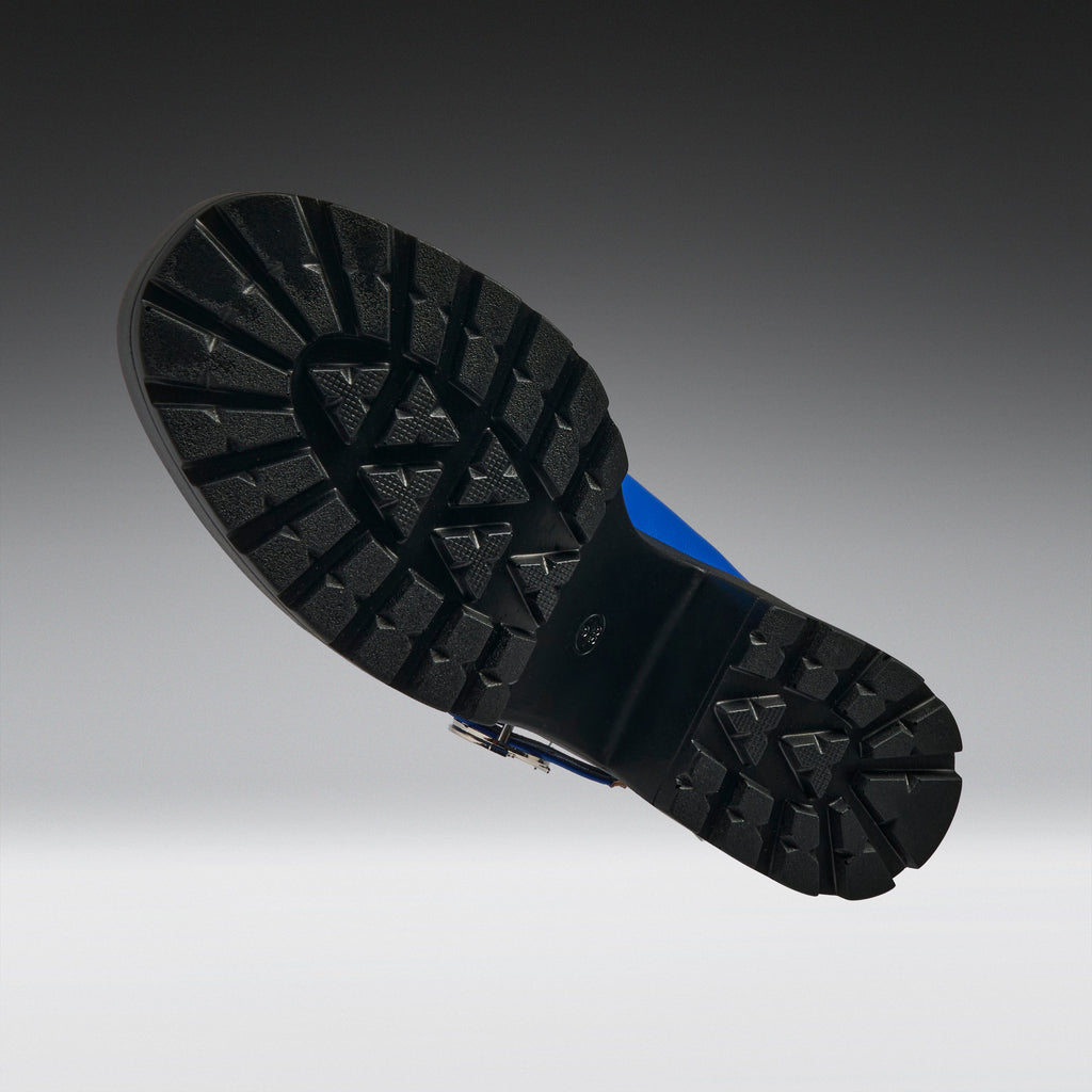 The treaded black rubber outsole of a boot, shown floating against a dark gray to light gray gradient background. A small blue accent is visible on the heel area of the sole.