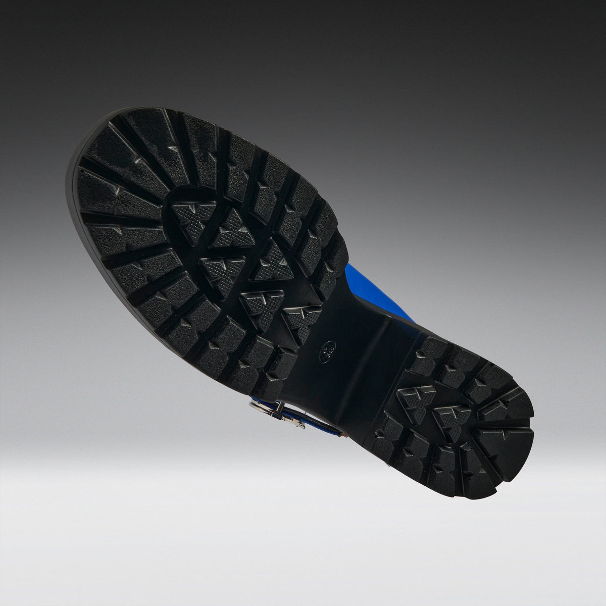 The treaded black rubber outsole of a boot, shown floating against a dark gray to light gray gradient background. A small blue accent is visible on the heel area of the sole.