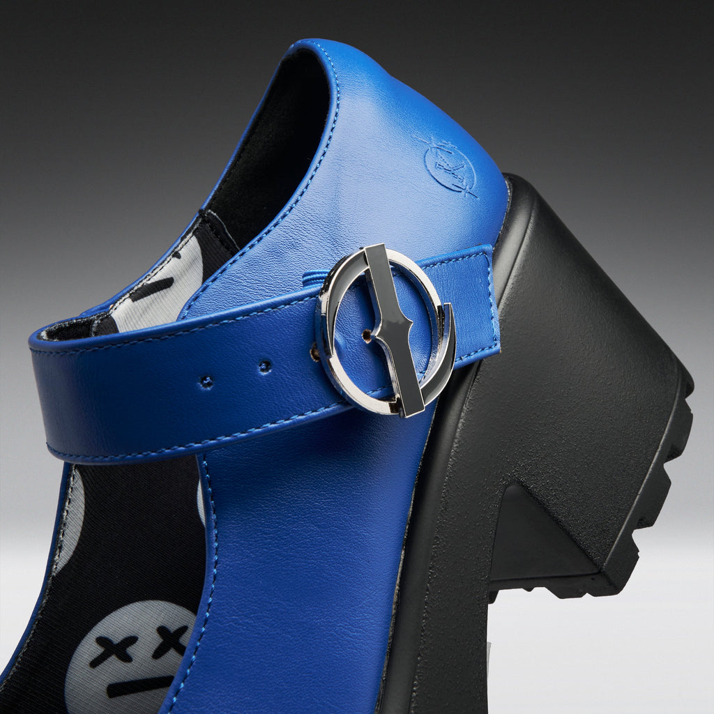 A close-up studio shot of a blue leather Mary Jane-style shoe with a chunky black platform heel and a silver buckle featuring a stylized 'X' emblem.