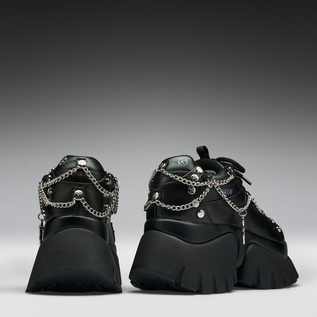 A pair of black chunky platform sneakers with silver chain and stud embellishments, shown from the back against a dark gray gradient background.