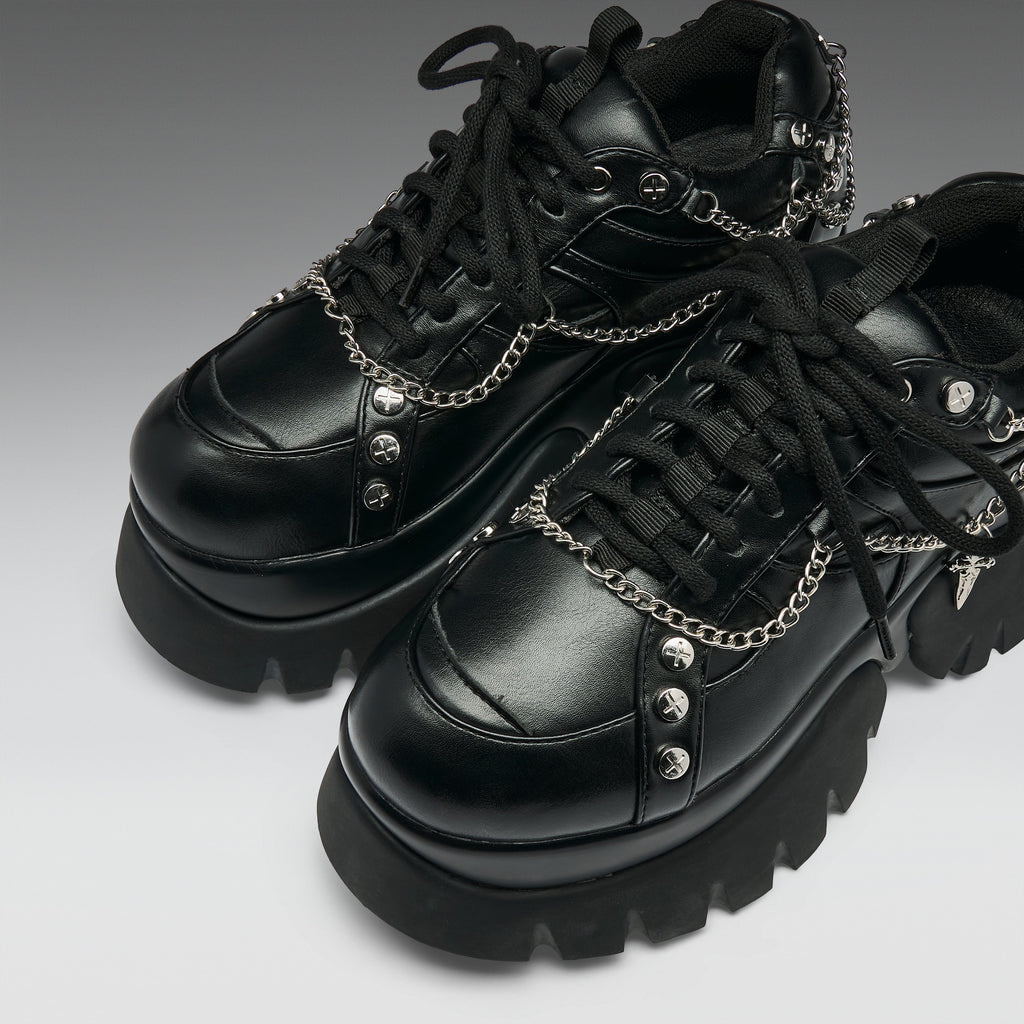 A pair of black leather platform sneakers with chunky lug soles, black laces, and silver chain details with skull and spike embellishments, shown on a gray gradient background.