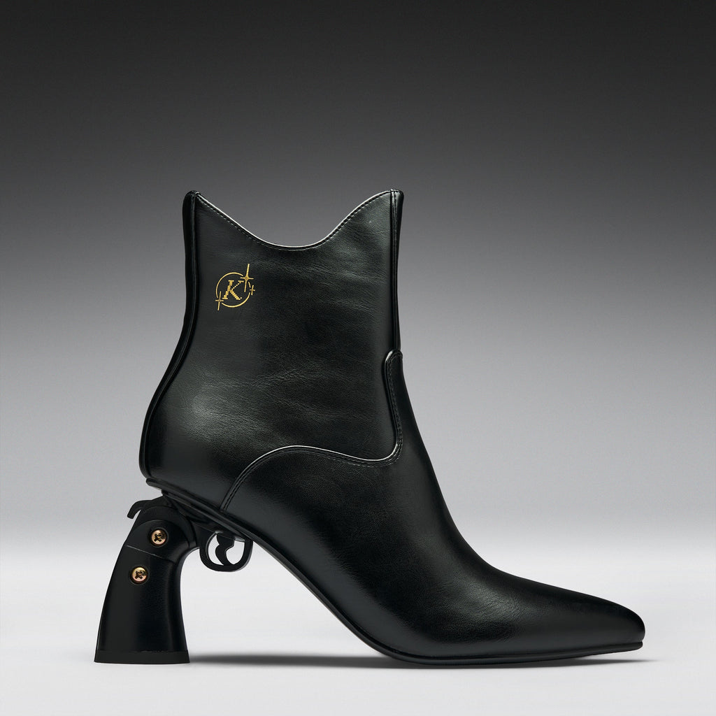A black leather ankle boot with a pointed toe, a unique pistol-shaped block heel, and a gold Karl Lagerfeld logo on the side, shown against a dark gray gradient background.