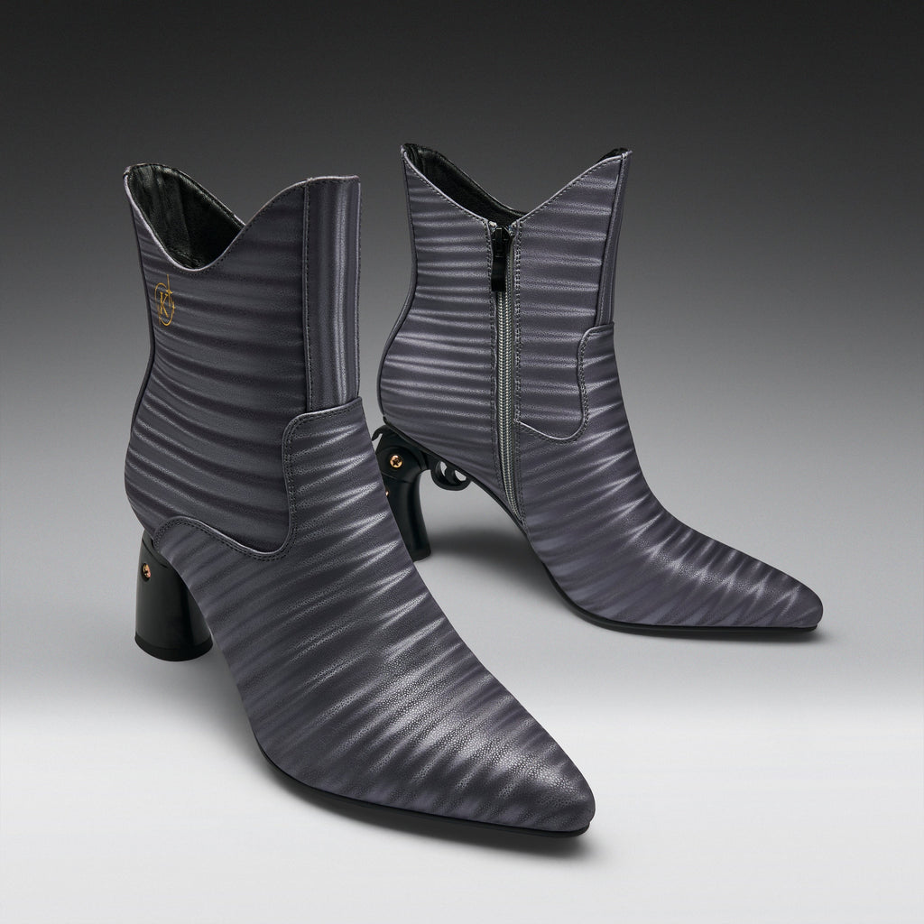 A pair of pointed-toe ankle boots in a dark gray ribbed leather, featuring a mid-height block heel, side zipper, and a small gold logo on the inner side, set against a dark-to-light gray gradient studio background.