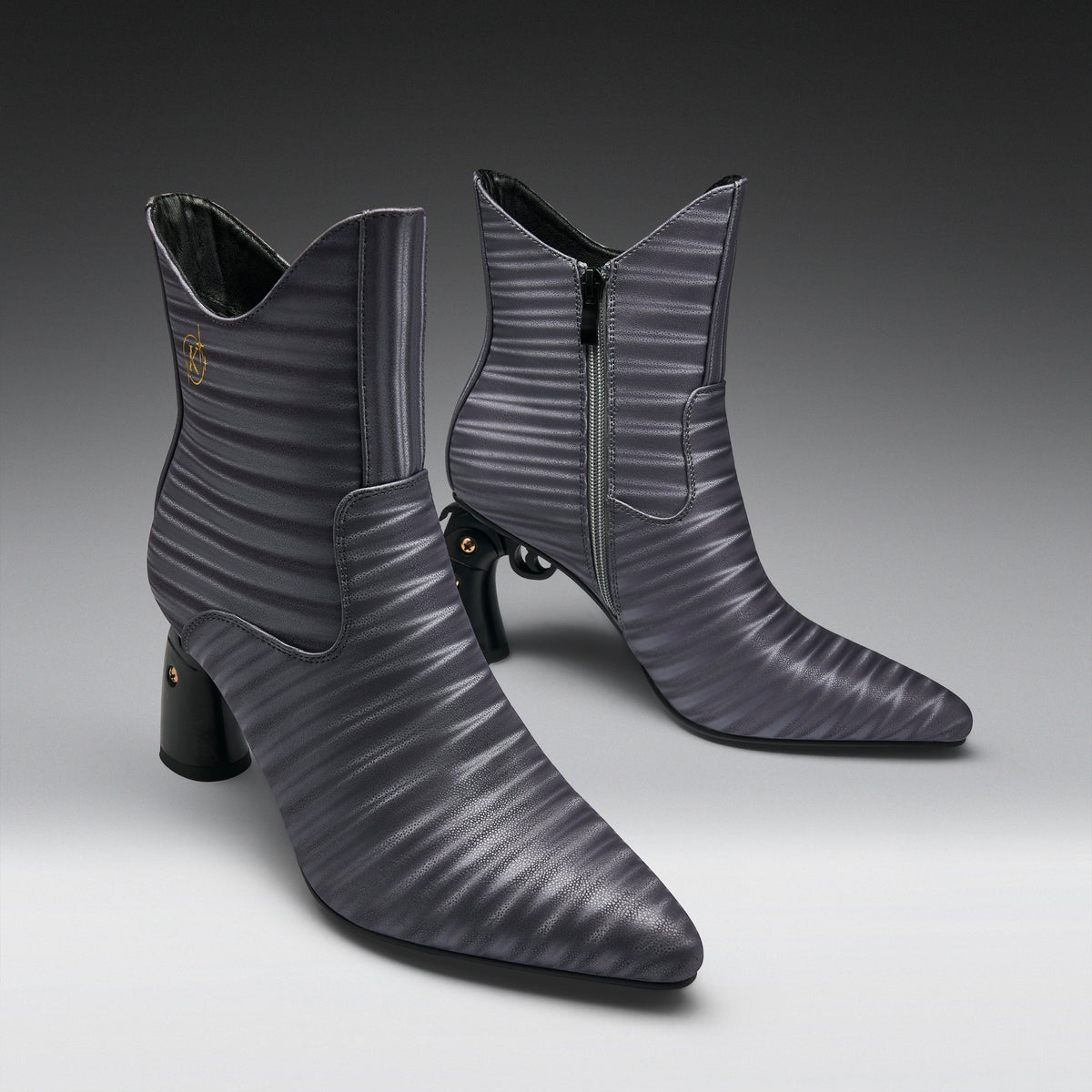 A pair of pointed-toe ankle boots in a dark gray ribbed leather, featuring a mid-height block heel, side zipper, and a small gold logo on the inner side, set against a dark-to-light gray gradient studio background.