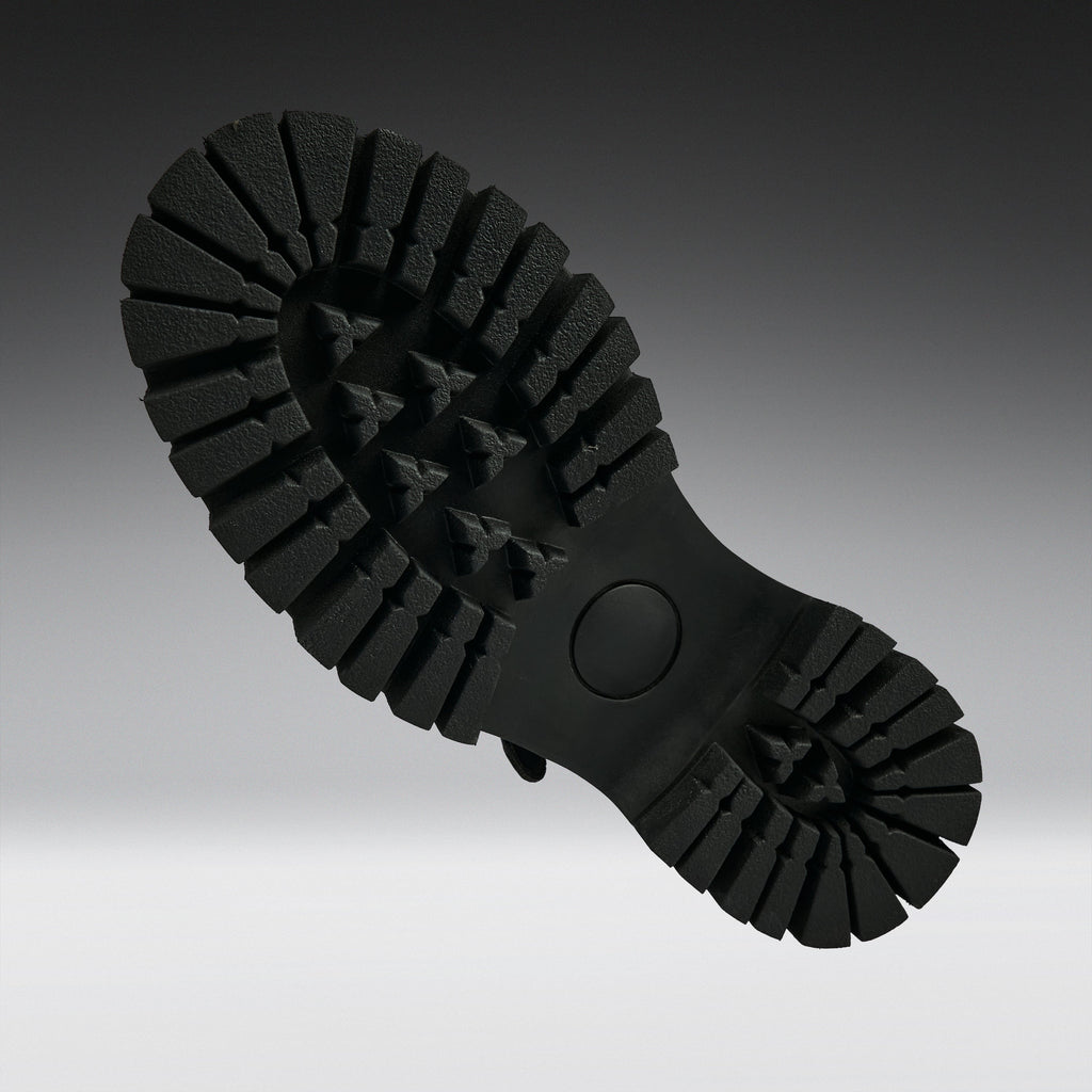 A close-up of a black rubber shoe sole, showing the deep lugs and tread pattern, photographed against a dark-to-light gray gradient background.