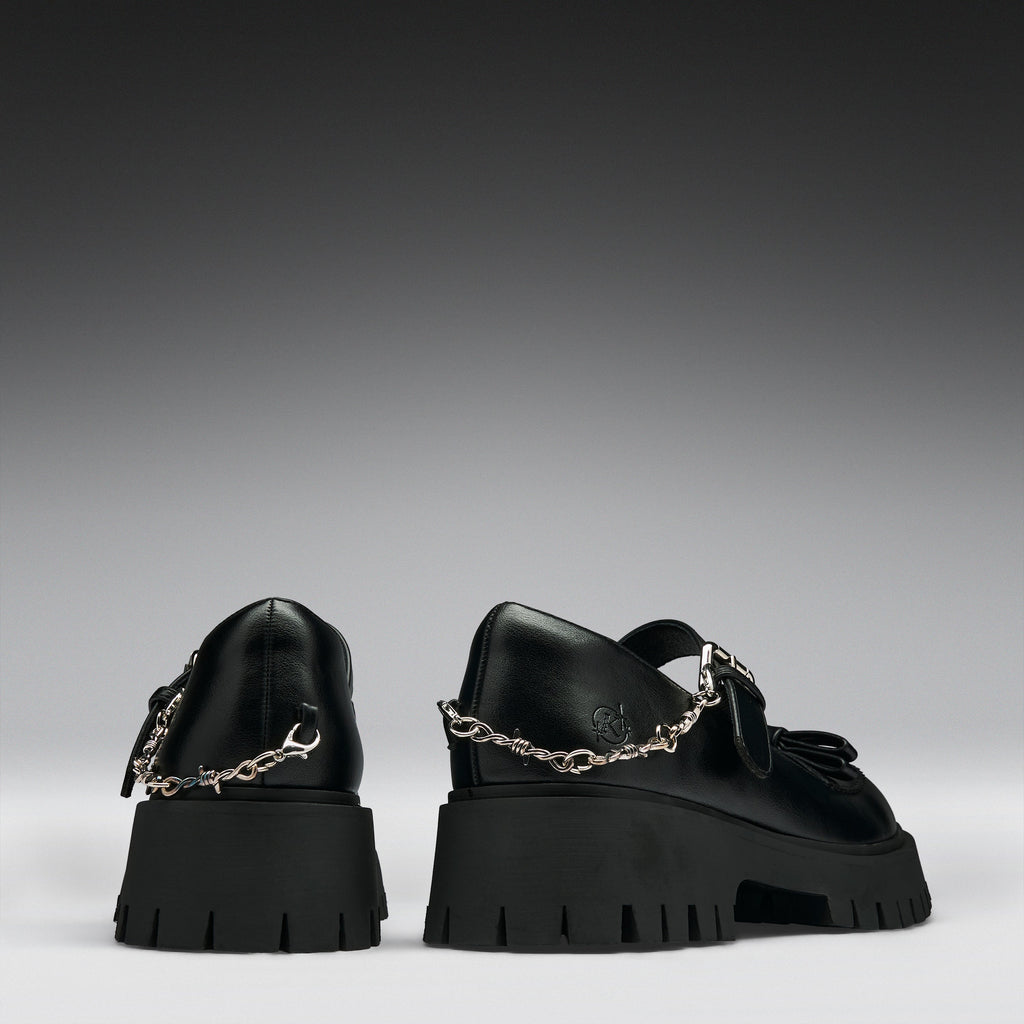 A pair of black leather Mary Jane shoes with chunky lug soles and silver chain details, shown from the back against a dark gray gradient background.