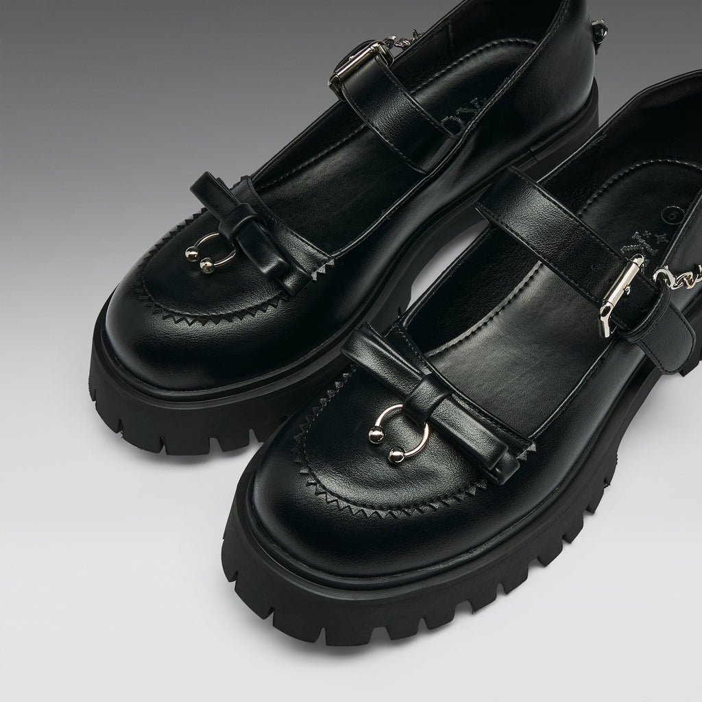 A pair of black leather Mary Jane-style shoes with chunky lug soles, decorative bow details, and silver-tone metal hardware, photographed on a clean gray studio background.